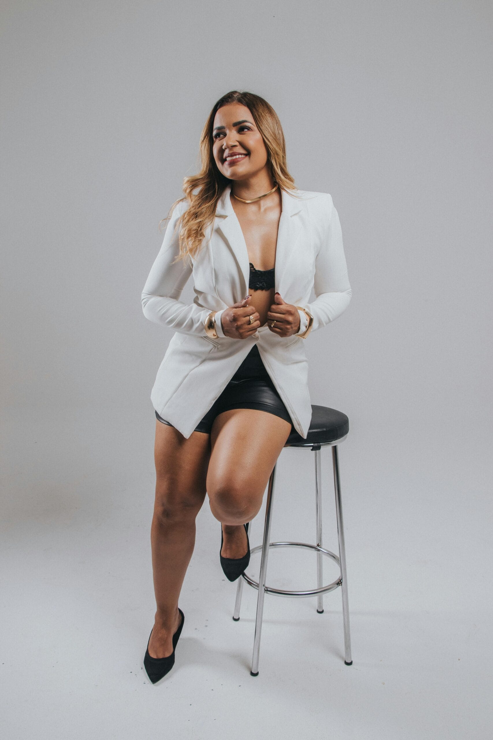 Smiling woman in white blazer sitting on stool in studio setting.